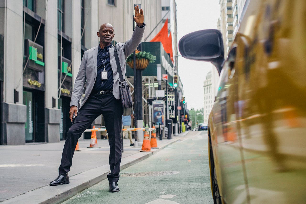 Full body of cheerful African American male with hand up catching bright yellow taxi on modern street of city